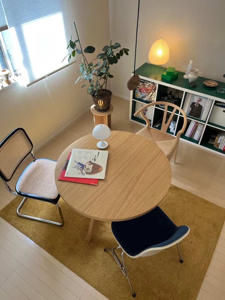 Light brown natural-toned studio living room featuring a round dining table, mixed chairs, plants, and warm lighting in a cozy atmosphere