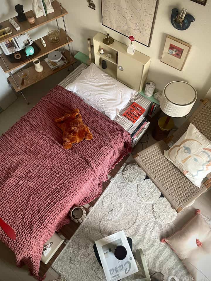 Warm natural-toned bedroom featuring wooden shelves, cozy bed with red blanket, and vintage table lamp