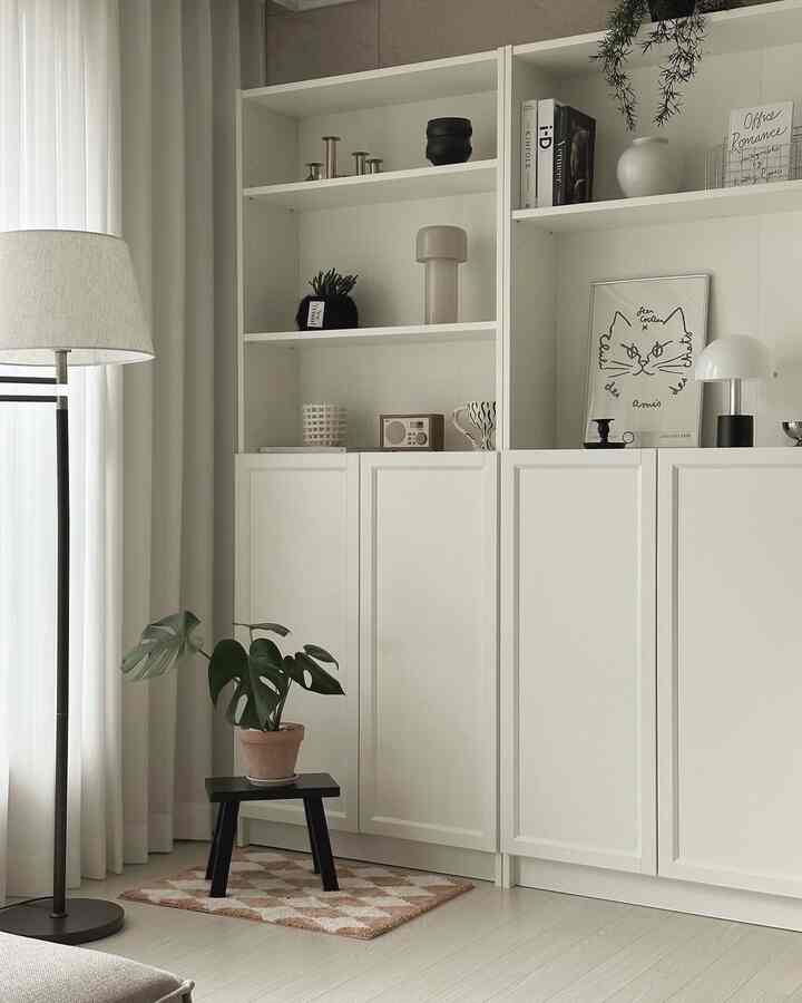 ホワイトとベージュトーンのリビング、ホワイトのBILLY本棚、フロアランプ、黒のスツールとテラコッタの植物が配置されたミニマルなインテリア