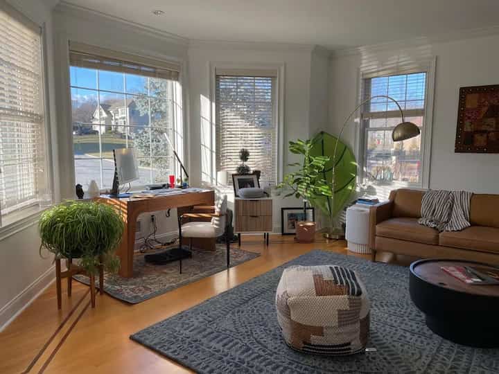 Bright living room filled with natural light features a wooden desk, brown leather sofa, and patterned pouf creating a cozy atmosphere