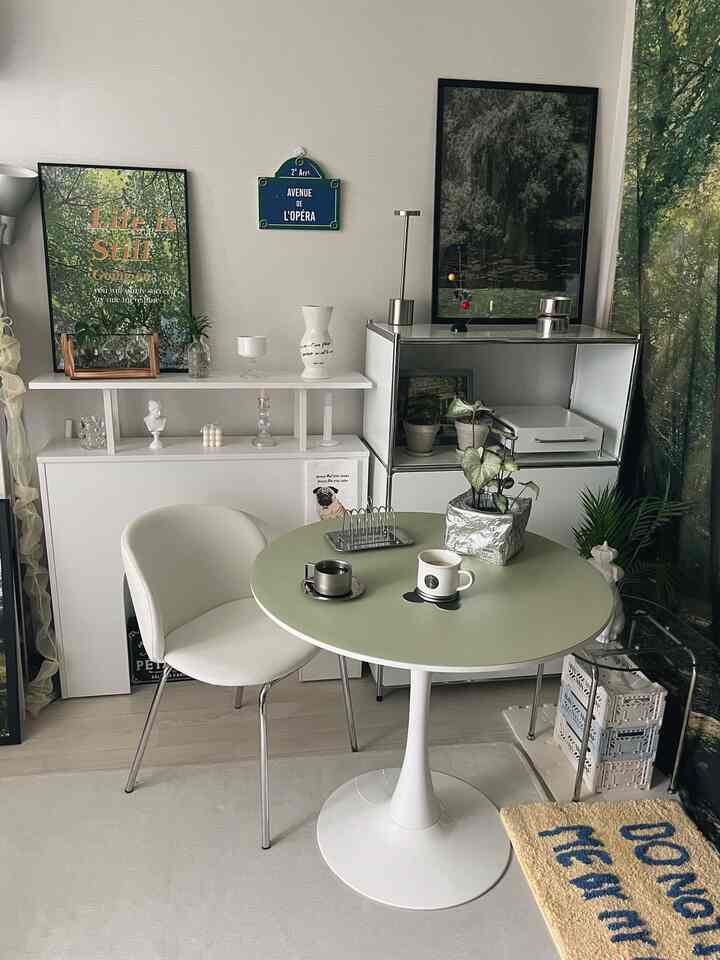 白と緑のトーンのリビング、シンプルなテーブルとチェア、額縁、収納棚、植物が配置された落ち着いた雰囲気のインテリア