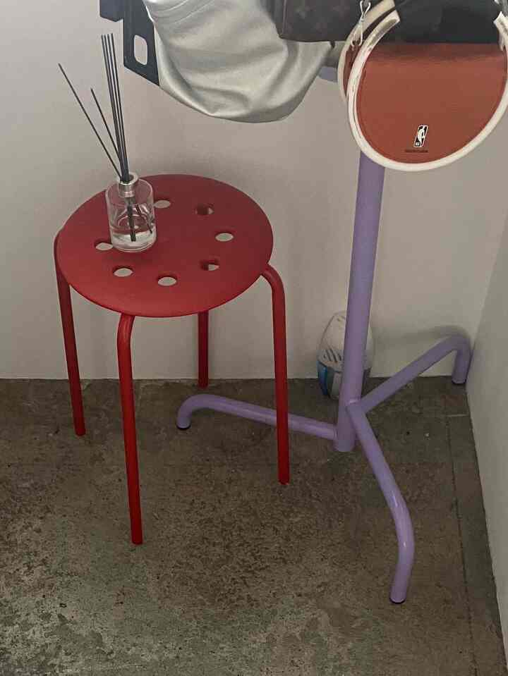 赤い鉄製スツールとラベンダー色のポールハンガーが置かれたシンプルなコーナーのインテリア