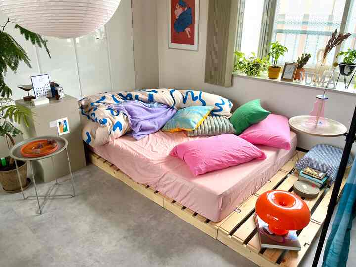 明るい自然光が差し込むナチュラルな色調の寝室、木製ベッドとカラフルな寝具が配置されたシンプルな雰囲気のインテリア