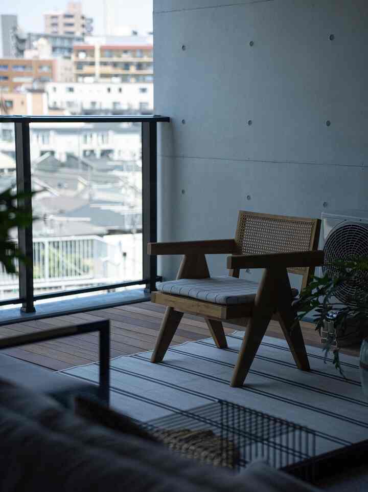 A modern and serene balcony with gray concrete wall, wood-tone armchair, and striped rug on wooden terrace flooring