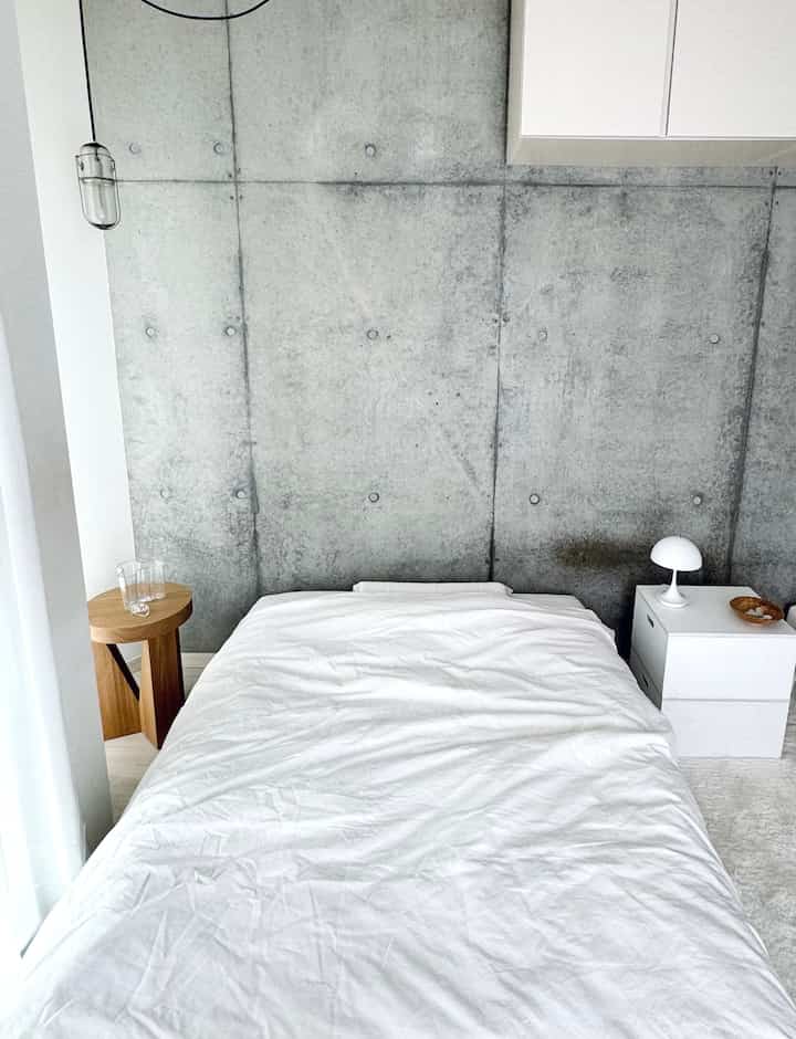 白とナチュラルトーンの寝室、コンクリートの壁とシンプルなベッド、右側のナイトテーブルにテーブルランプが置かれたモダンなインテリア