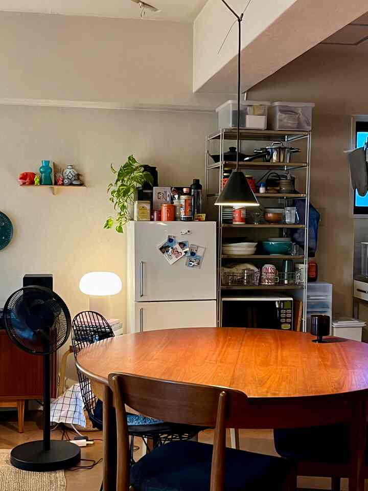 ブラウンの木製丸テーブルと椅子が配置されたダイニングルーム、白い冷蔵庫と金属製の棚がある雰囲気のあるキッチン空間