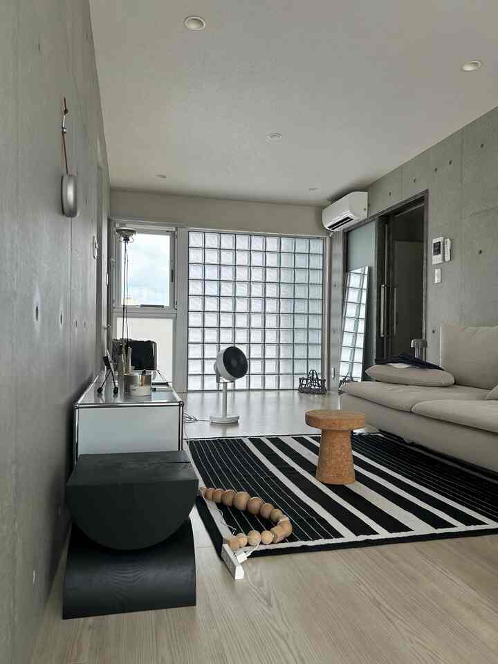 A beige and black toned living room featuring a beige sofa, striped rug, cork side table, and a circulator fan in a modern natural style
