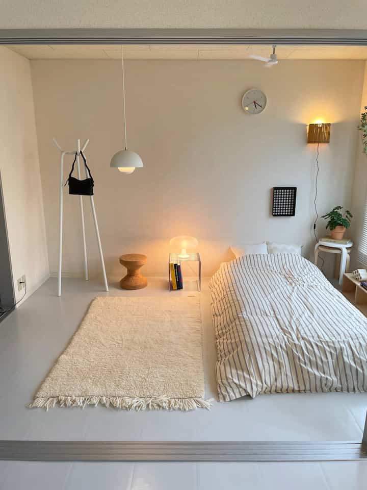 白とナチュラルカラーの寝室、ロータイプのベッドとコルクスツール、ペンダントライトと壁灯が配置された落ち着いたインテリア