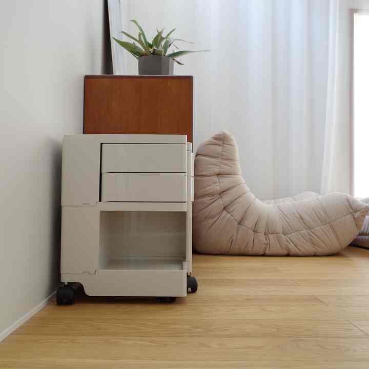 ナチュラルな木の床と白い壁があるシンプルなリビング、白いボビーワゴン収納とベージュのビーズクッションが配置された印象的な空間