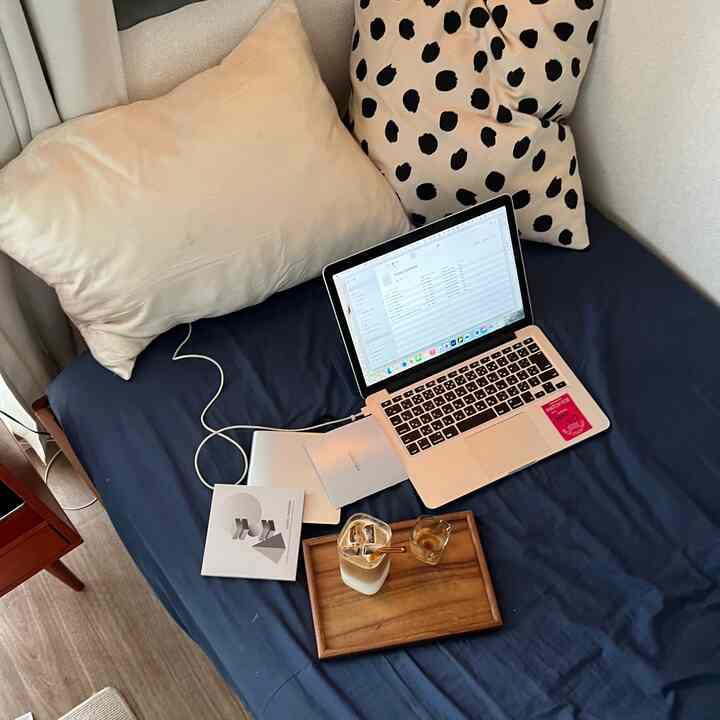 An 8畳 bedroom with navy bed sheets and gray cushions, featuring a laptop and wooden tray with iced coffee in a cozy single household space