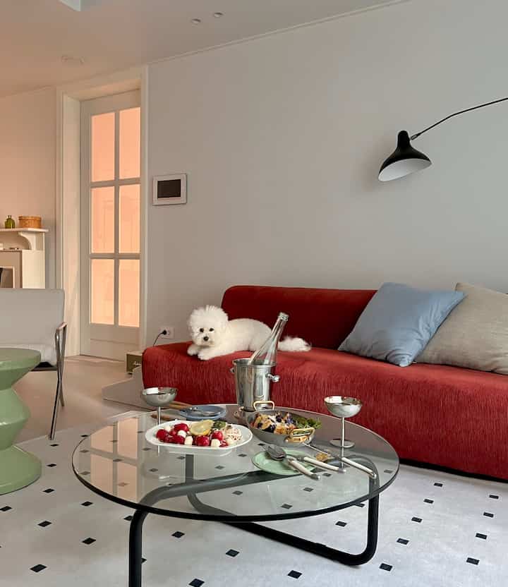 Living room with white walls and a red sofa, featuring a glass coffee table with wine glasses for a cozy home party setup