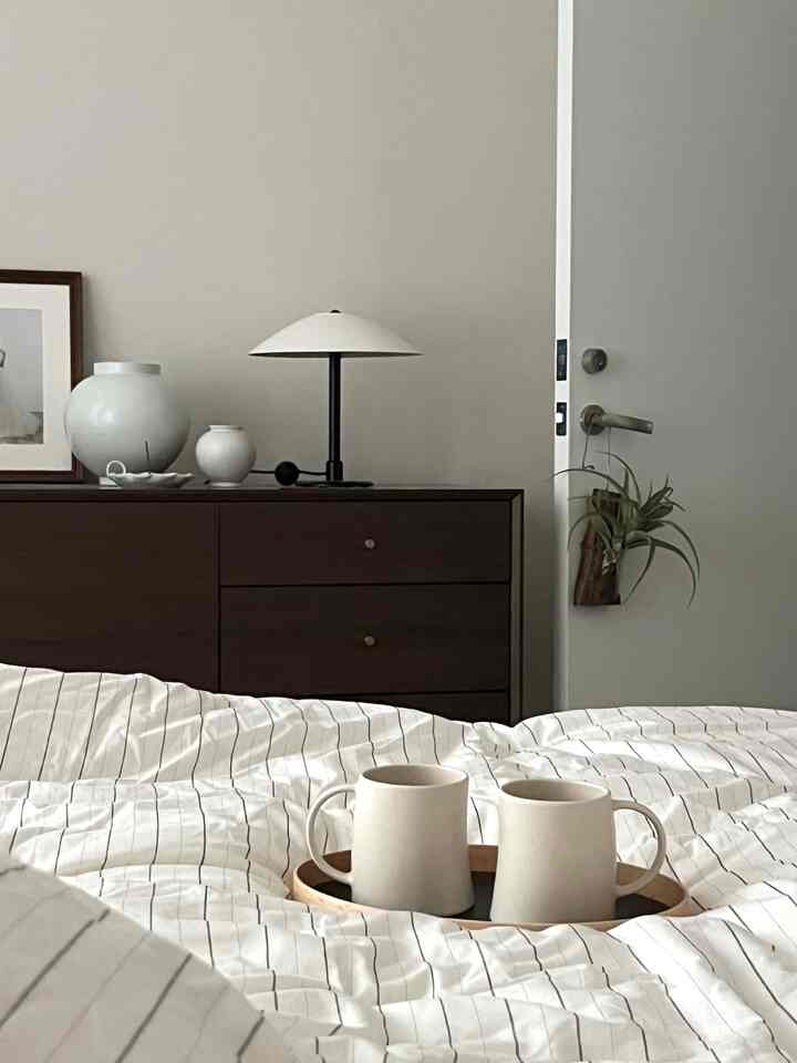 Natural modern bedroom featuring a bed with striped bedding, brown dresser, table lamp, and mugs on a tray creating a cozy atmosphere