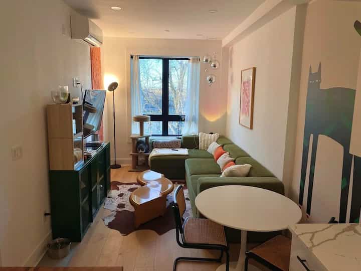 Modern minimalist living room with a green sectional sofa and round dining table, featuring a cat tower next to the window