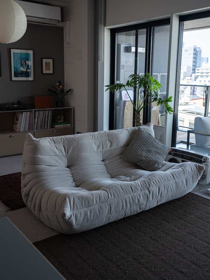 ナチュラルトーンのリビングルーム、中央に低く柔らかなロゼトーゴソファ、大きな窓際に緑の植物が配置された雰囲気のあるインテリア