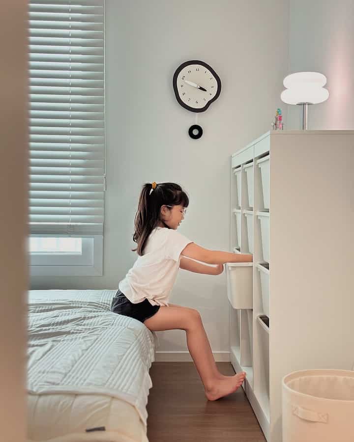 明るいホワイト調の寝室、窓際にベッド、右側の壁に収納が配置され、子供が触れている雰囲気のあるインテリア