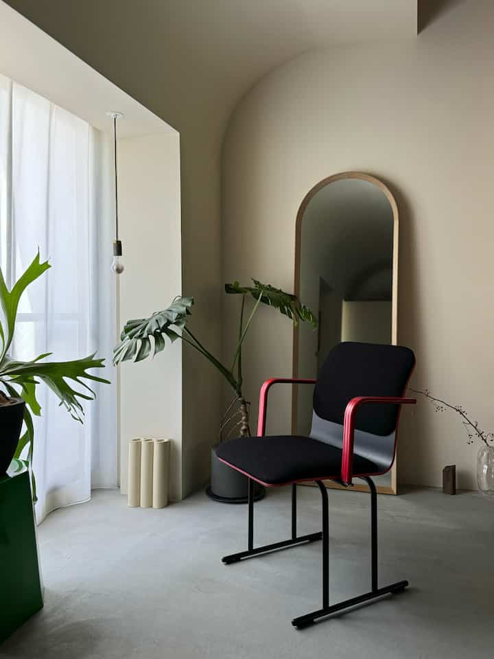 Natural beige walls and gray concrete floor in a living room, featuring a vintage stool with red armrests and black cushion, complemented by a large green plant in a minimal atmosphere