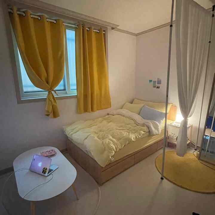 Yellow-toned studio apartment bedroom featuring yellow curtains, a bed, and a small coffee table arranged in a cozy compact space