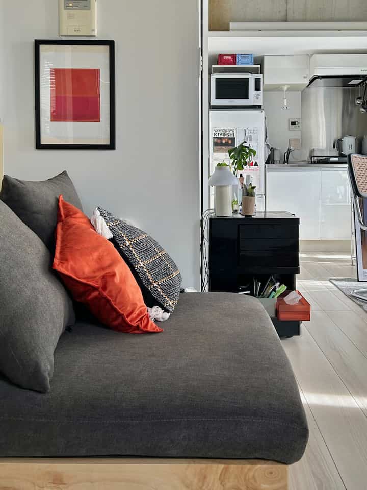 グレーのトーンが印象的なワンルームのリビング、ローソファと赤いクッション、キッチンが見えるシンプルでモダンなインテリア