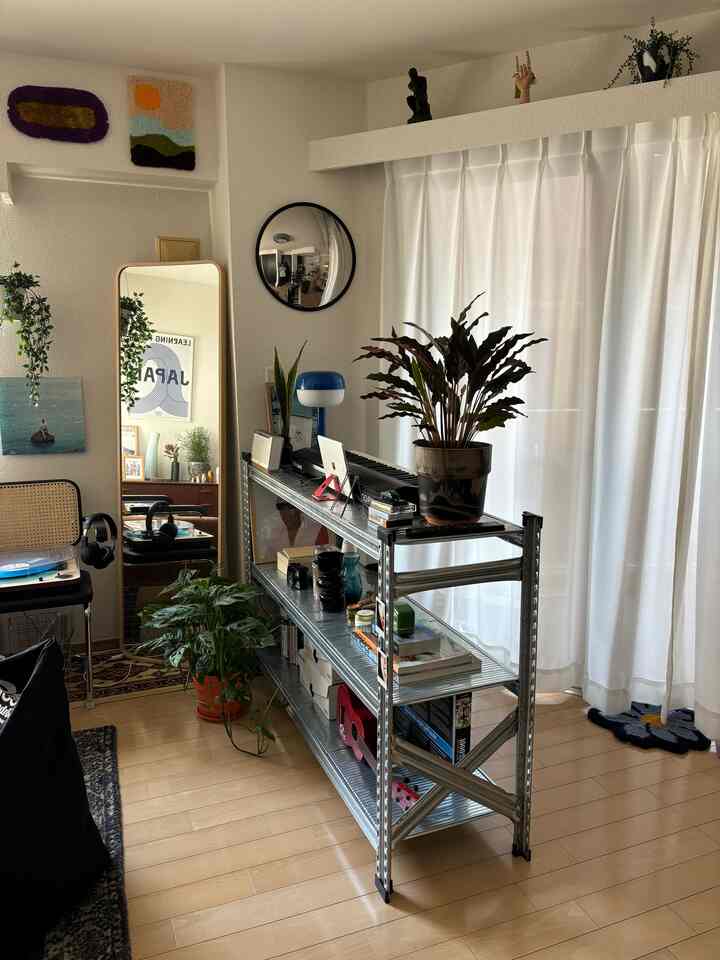 Bright living room with white curtains and wood tone flooring featuring an open metal rack and lush foliage plants creating a natural vibe.