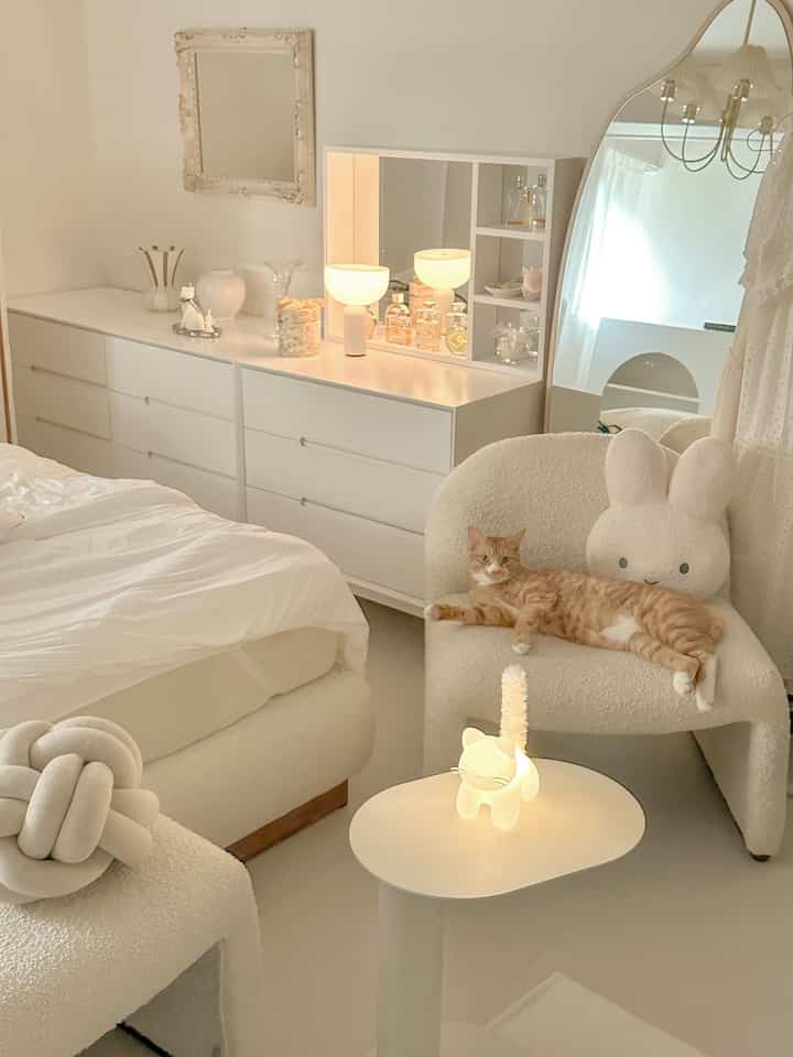 柔らかいホワイトトーンの寝室で、くつろぐ猫がいるアームチェアとかわいいムードランプが配置された雰囲気のあるインテリア