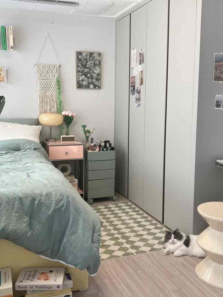 淡いグリーンとベージュの色調の寝室、寝具、ナイトテーブル、ラグが配置されたナチュラルな雰囲気のインテリアで猫が床に横たわっている