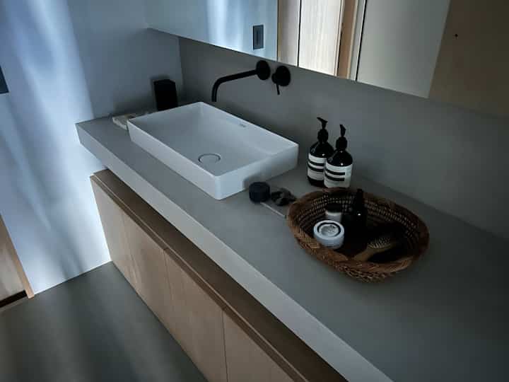 グレーとナチュラルウッドのバスルーム化粧台、白い洗面台とブラック蛇口が配置され、アロマ製品が整然と並ぶ雰囲気のあるインテリア