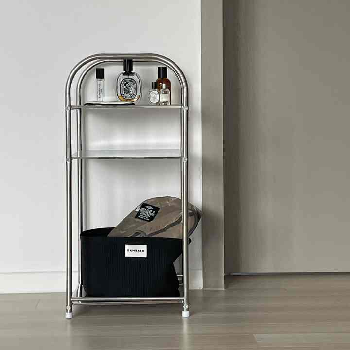 グレーとホワイトトーンの玄関、メタルフレームの棚に香水が並び、下には黒いファブリックバスケットが置かれた清潔感のある空間