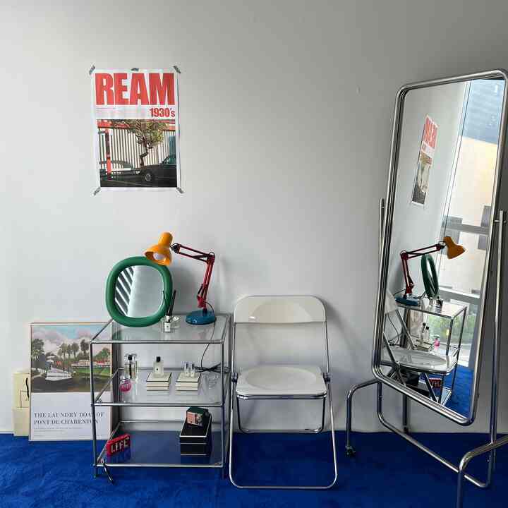 ホワイトの壁とブルーカーペットのホームオフィス空間、シルバーフレームの鏡と棚、オレンジのテーブルランプと白い椅子が配置されたミッドセンチュリーモダンのインテリア