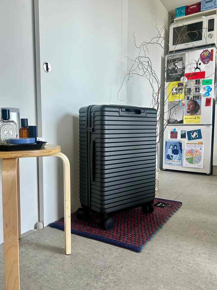 Minimalistic studio entrance with a black suitcase, wooden stool, gray floor, and white walls creating a neat atmosphere