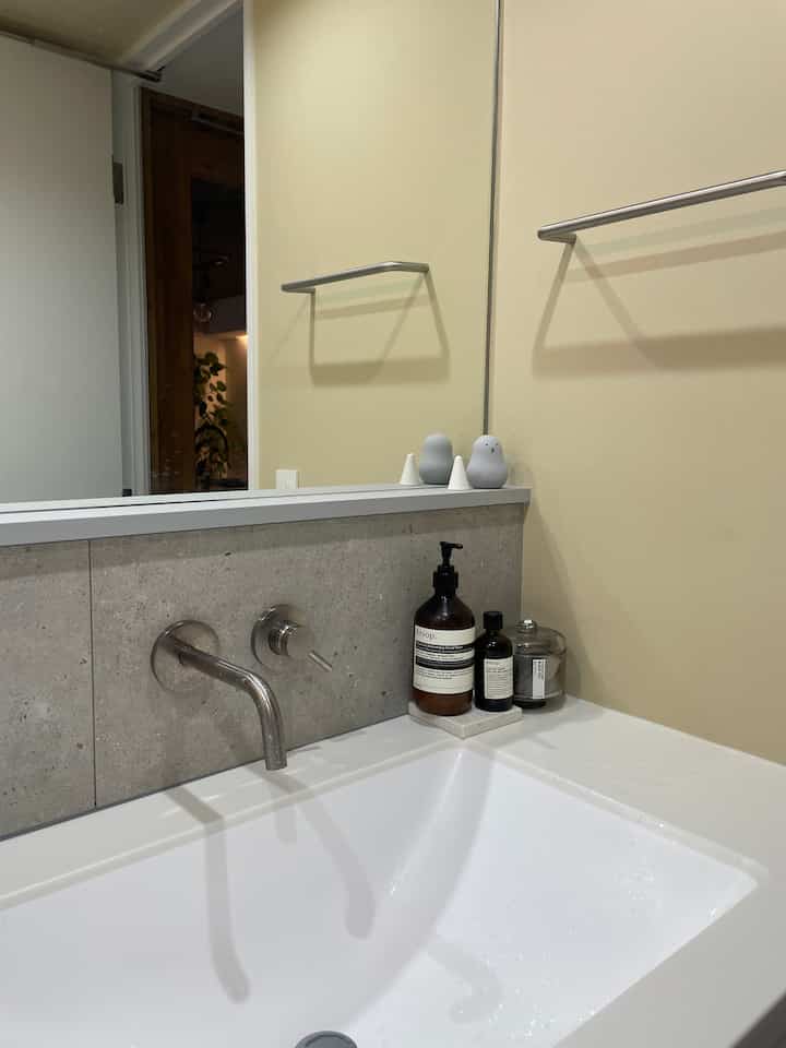 ベージュの壁と白い洗面台が配置されたモダンでシンプルなバスルーム空間、洗練されたクローム製壁付け混合水栓とタオル掛けが特徴的なインテリア