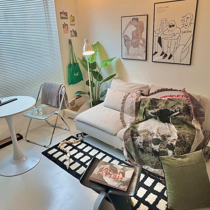 白と緑の落ち着いた色調のコンパクトなリビング、ソファとスツール、植物、ブラインドが配置された雰囲気のある空間