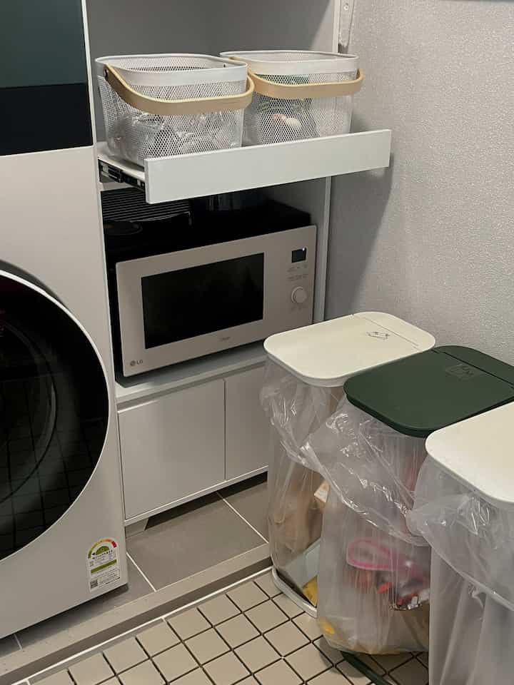 白とベージュのトーンのキッチン多目的室、整理された分別ゴミ箱と電子レンジ収納キャビネットが配置された実用的な空間