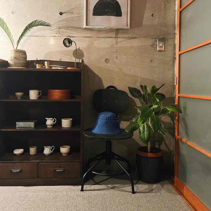 Natural-toned corner space with concrete walls, featuring a unique black chair with a blue hat, plants, and wooden shelving creating a cozy atmosphere