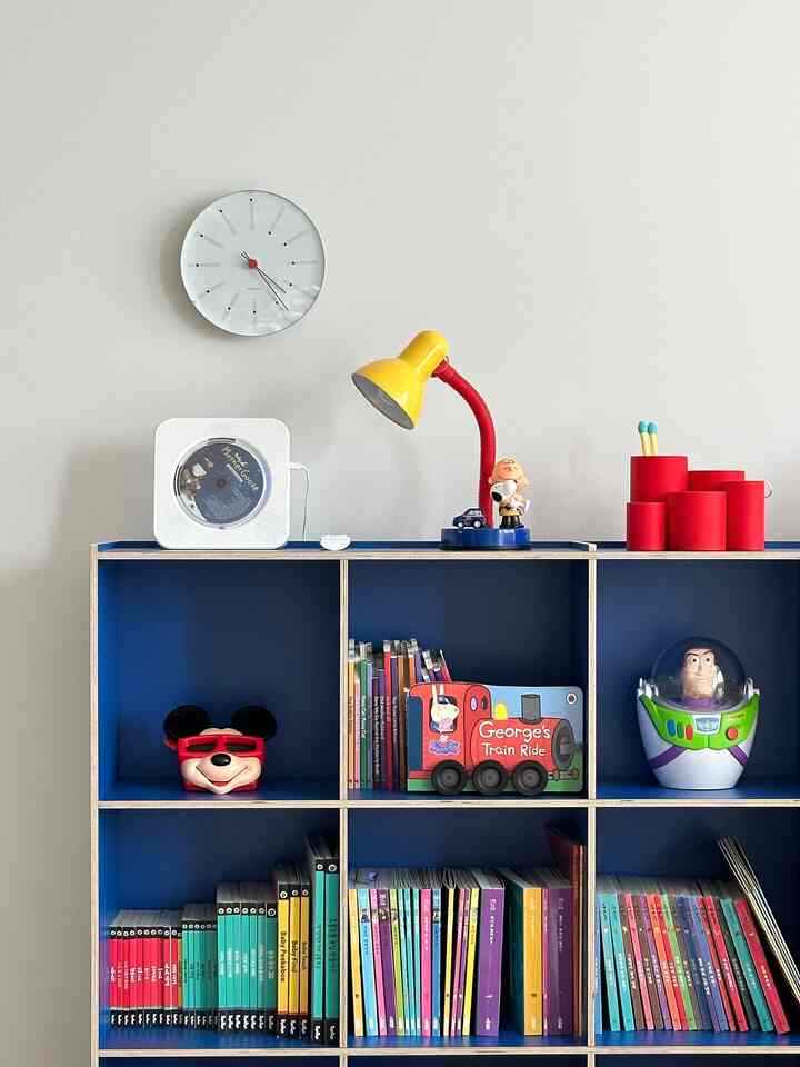 青い本棚に色とりどりの絵本やおもちゃが並ぶ子供部屋、白い壁と赤いデスクランプが印象的な明るくかわいらしいインテリア
