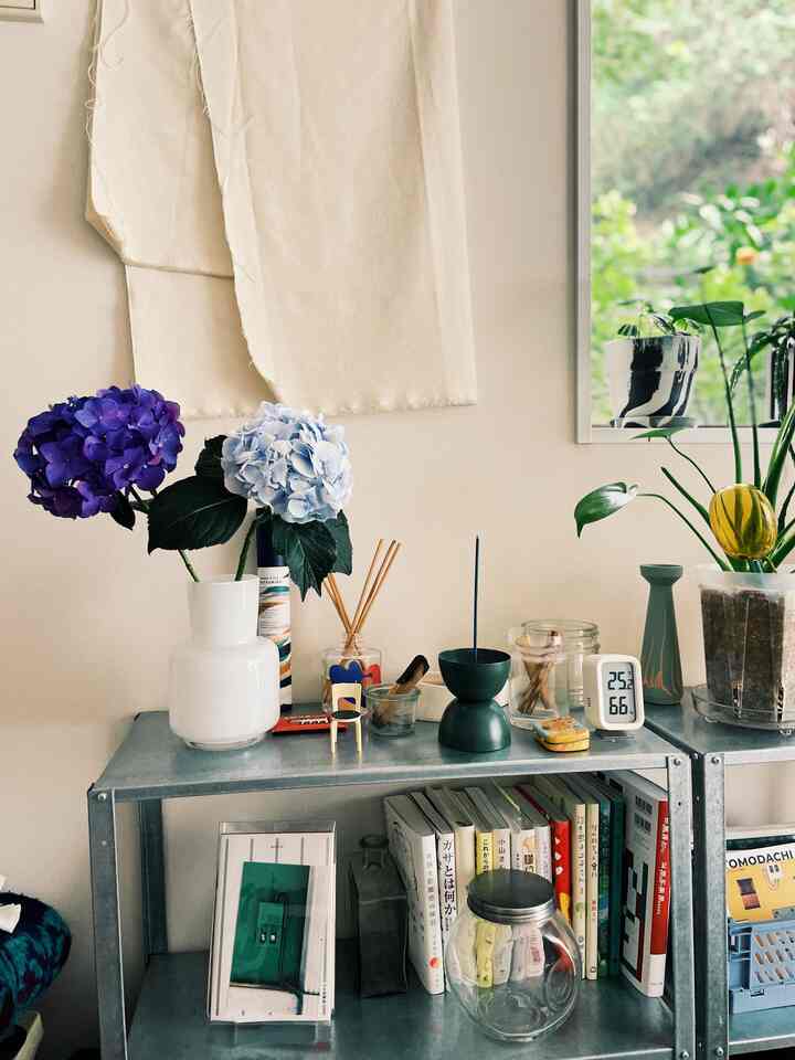 Beige-toned wall and small metal shelving unit with purple and light blue hydrangeas in a vase and various plants arranged in a neat single household space