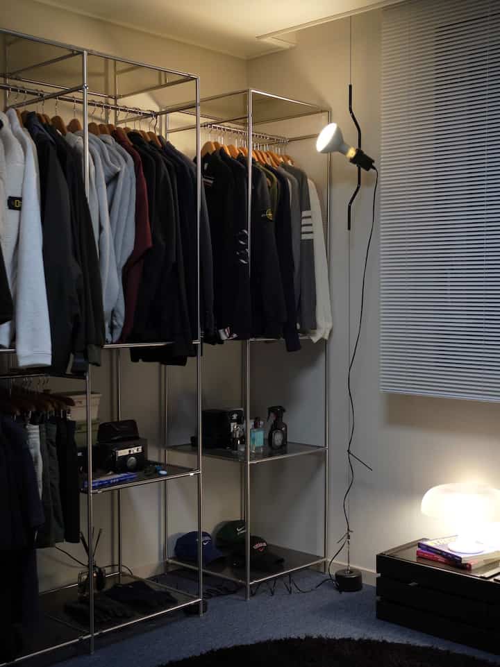 メタルフレームのウォークインクローゼットにネイビーと黒の衣類が並び、白いブラインドとペンダントライトが印象的なモダンなインテリア