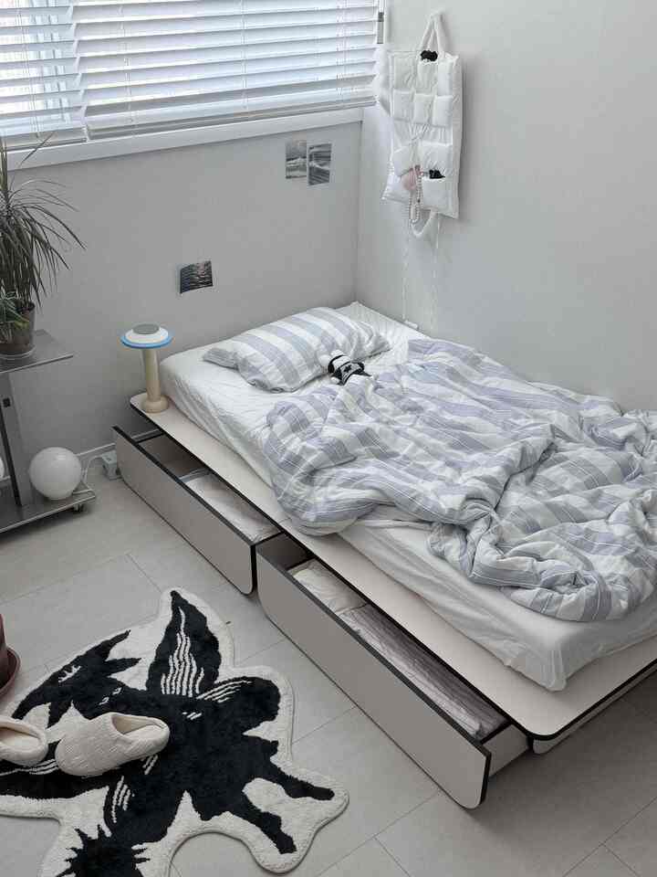 白と木目調が調和した小さな寝室、収納付きベッドとブラインドが配置されたシンプルで居心地の良い空間