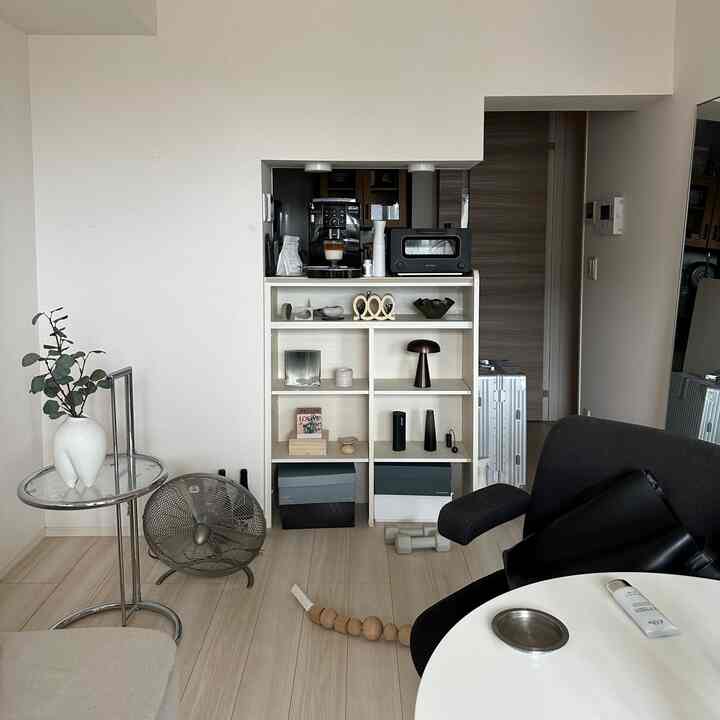 A modern natural living room with white walls and wood-tone flooring, featuring central shelves with a coffee machine and decor accessories