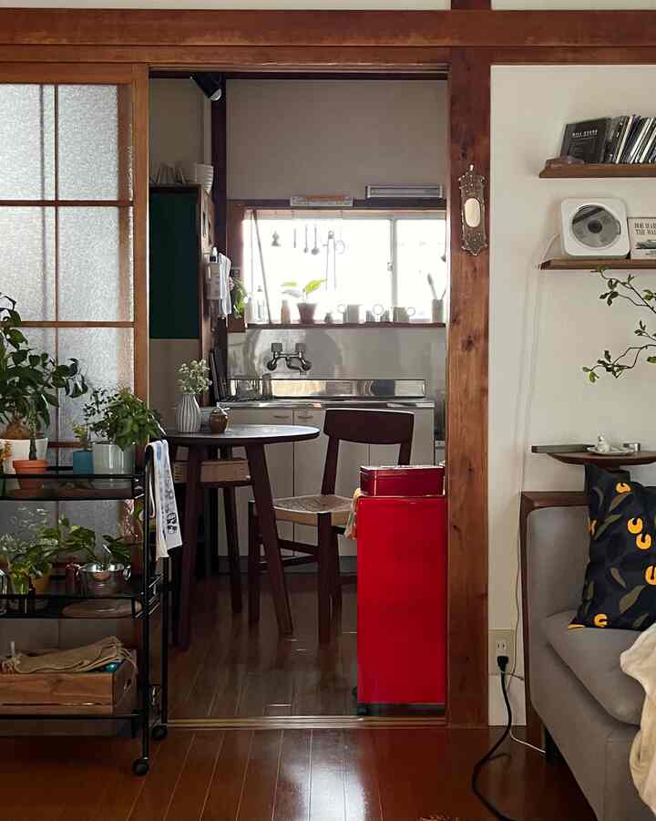 木目調と赤のアクセントが調和するワンルームのキッチンとリビングスペース、ダイニングテーブルと植物が配置された温かみのある雰囲気のインテリア