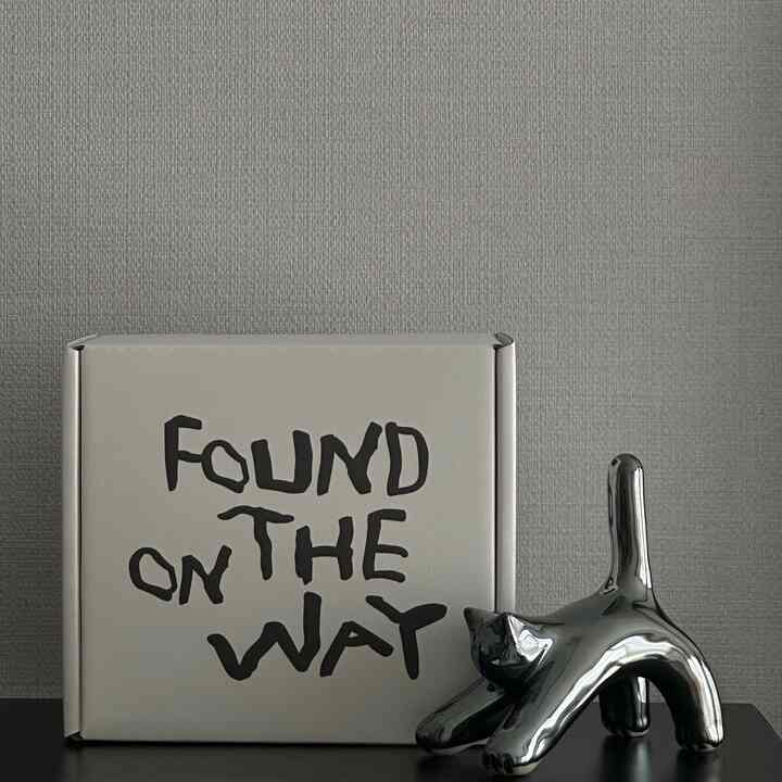 グレーの壁の前に黒い棚の上に白い箱と銀色の猫のオブジェが置かれたミニマルなインテリア空間