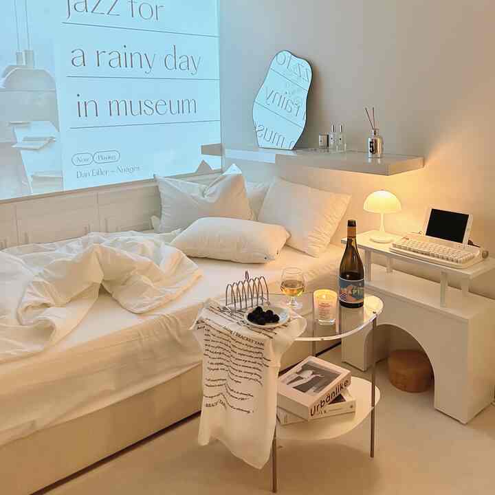 Bright white-toned bedroom featuring a bed, wave-shaped mirror, and wireless table lamp in a minimal, cozy atmosphere