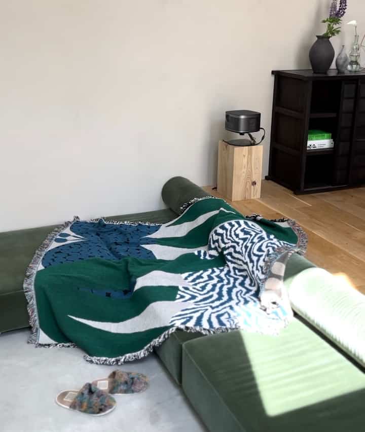 ナチュラルなグリーントーンのリビング、緑のセクショナルソファにカラフルなスローと木目調の床が調和した雰囲気のある空間