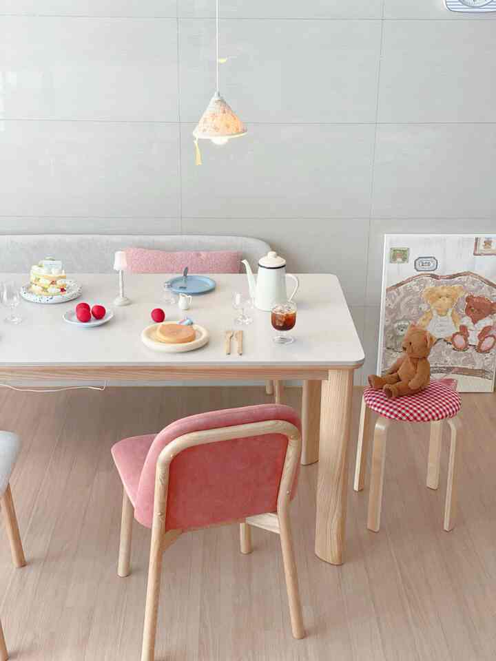 White-toned dining room featuring a wooden dining table, pink dining chairs, and a teddy bear creating a cozy atmosphere
