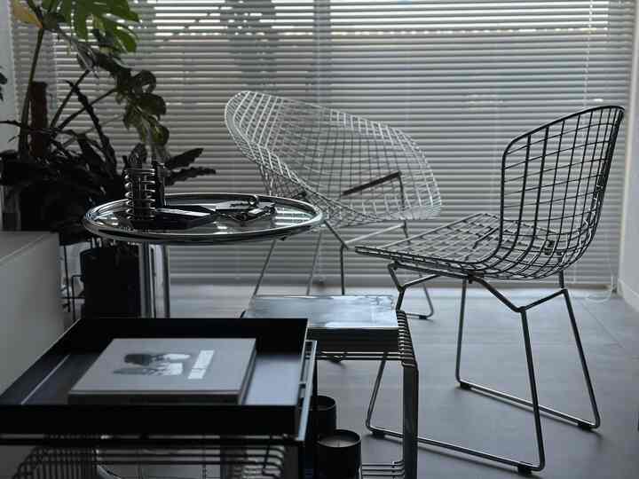 白いブラインドの背景に金属製ワイヤーチェアとガラスのサイドテーブルが配置されたモダンなリビングスペース
