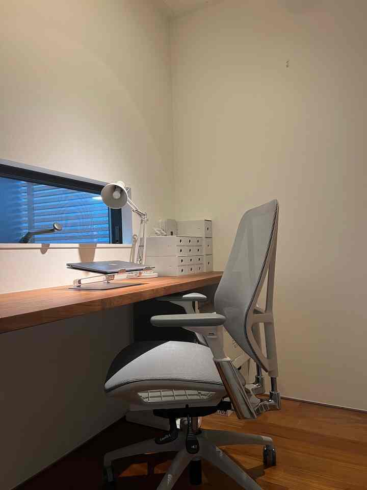 Monotone home office with white walls and wood tone desk, featuring ergonomic office chair in a compact space