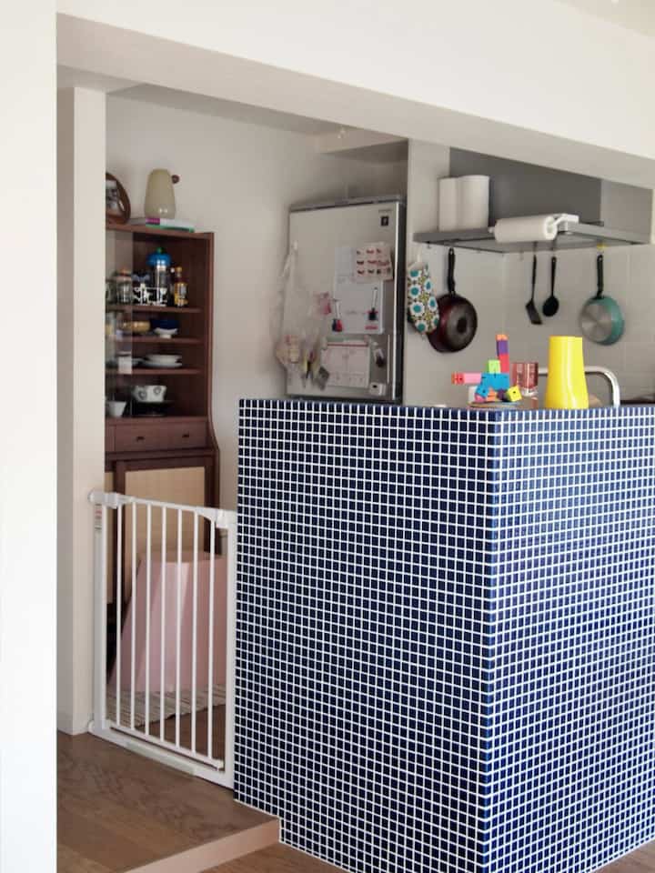 白い壁と青いタイルのキッチンカウンターがあるキッチン空間、左側に赤ちゃんの安全のためのベビーゲートが設置された温かみのある雰囲気のインテリア