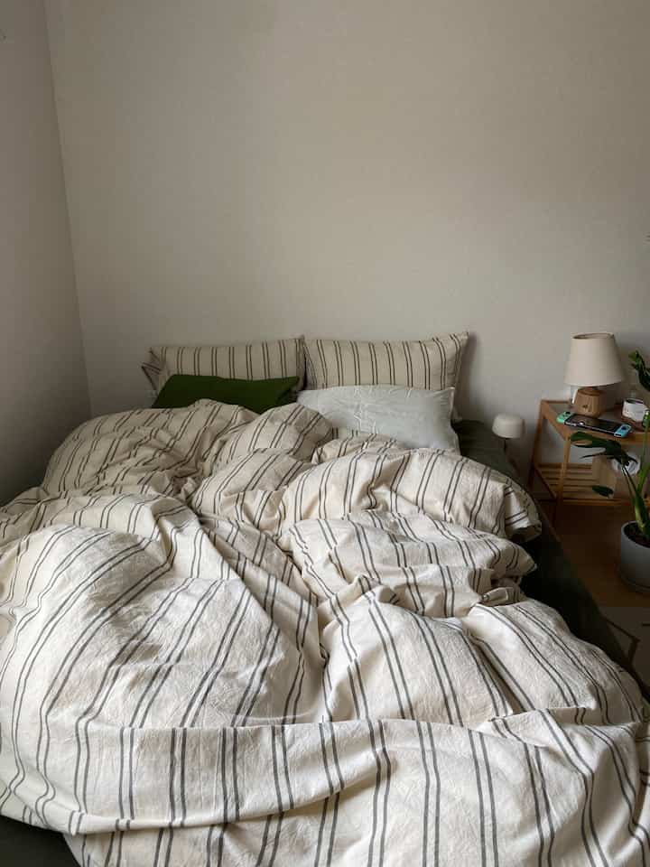 白とベージュの色調の寝室、ストライプの寝具とナイトテーブル、テーブルランプが配置された雰囲気のあるインテリア