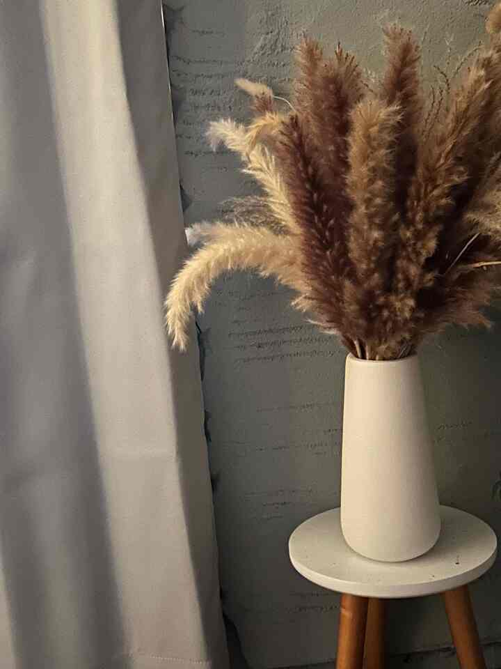 Beige and white toned bedroom corner featuring a wooden stool with a ceramic vase holding dried pampas grass, creating a natural ambiance