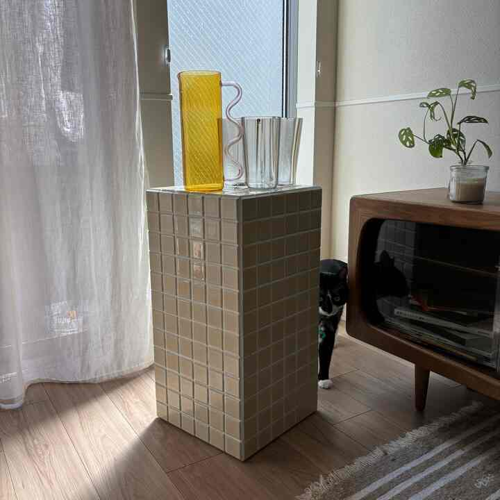 Natural-toned living room with beige cream tile stool, wooden TV stand, small plant, and a black cat peeking from behind