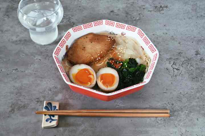 グレーのコンクリートテーブルの上に、赤い八角形のラーメン鉢に卵、肉、野菜を盛り付けたカフェ風の食事シーン